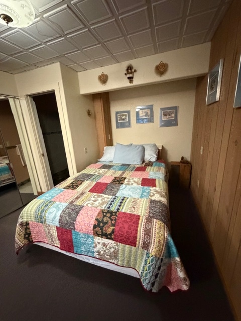 A queen bed with a colorful patchwork quilt, wood-paneled walls, and a white grid-patterned ceiling.