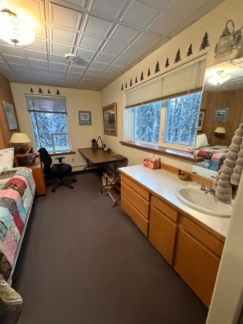 An elongated room featuring a wood-toned vanity with a white sink on the right, a desk with a black office chair in the background, and a bed with a colorful patchwork quilt on the left.