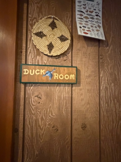 A close-up of a wood-paneled wall featuring a wooden sign that reads "DUCK ROOM" with an illustration of a duck. Above the sign hangs a circular woven basket with a dark diamond pattern, and a chart illustrating various fish species is pinned to the right.