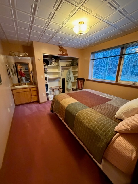A bedroom featuring a bed with a brown and green checkered quilt, a white grid-patterned ceiling with a central light fixture, and an open closet with shelving. To the left, a wooden vanity with a white sink and mirror is built into the peach-colored wall.
