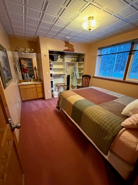 A bedroom featuring a bed with a brown and green checkered quilt, a white grid-patterned ceiling with a central light fixture, and an open closet with shelving. To the left, a wooden vanity with a white sink and mirror is built into the peach-colored wall.