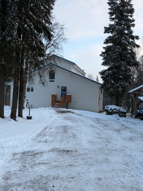 An exterior view of a white, two-story building in a snowy landscape, surrounded by tall, frost-covered evergreen trees. A small wooden deck with stairs leads to an entry door on the side of the house.