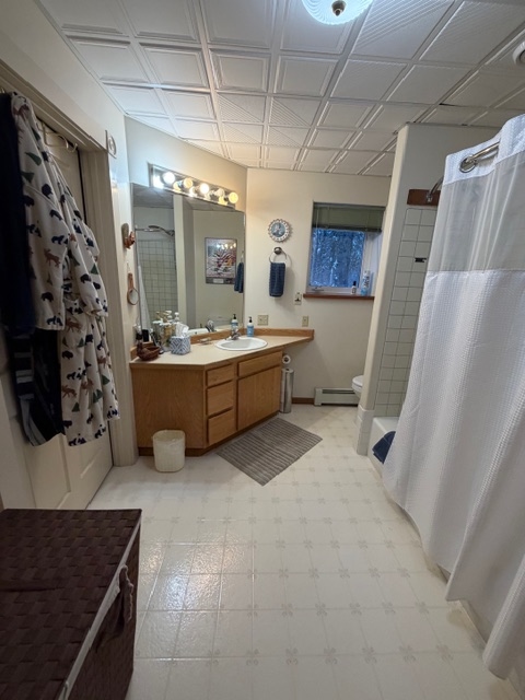 A spacious bathroom with white tiled flooring and a white grid-patterned ceiling. On the left is a wooden vanity with a white countertop and large mirror illuminated by a row of vanity lights. To the right, a white shower curtain is partially drawn, and a small window in the background provides a view of a snowy outdoor scene.