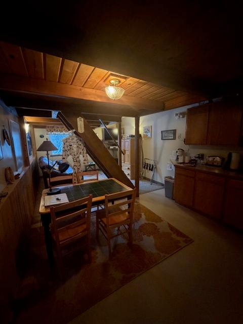A dimly lit interior space with wood-beamed ceilings and wood-paneled walls. In the foreground, a small wooden table with two chairs sits on a patterned rug. To the right, a kitchen area with dark wooden cabinetry is visible. In the background, a staircase leads upward, and a peek into a living area reveals a lamp and a window.