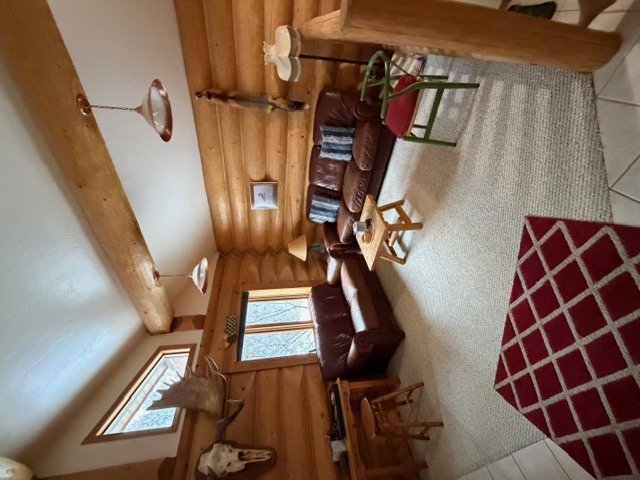 An interior view of a rustic living area featuring natural log walls and a white area rug with a small red and white diamond-patterned rug in the corner. The space is furnished with two brown leather sofas and a light-colored wooden coffee table. Decorative elements include a mounted moose head above a window, several animal skins on the walls, and a large exposed wooden log beam across the vaulted ceiling.