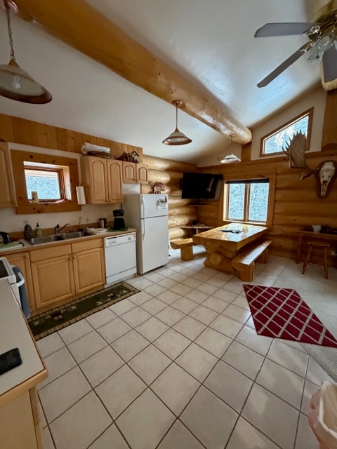 An interior view of a rustic kitchen and dining area with light-tiled floors and large exposed log beams on a vaulted ceiling. The kitchen features wooden cabinets, a white refrigerator, and a white dishwasher, while the dining area includes a large wooden picnic-style table with benches and a mounted moose head on the wall.