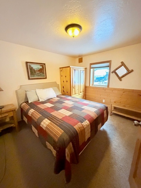 A bedroom featuring a bed with a warm, reddish-brown patchwork quilt and a light-colored headboard against a cream-colored wall. The room includes a window overlooking a snowy landscape, wood-paneled accents on the lower half of the walls, and a matching wooden armoire and nightstand.