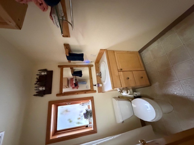 A bathroom featuring a light-colored vanity with a white sink and a wooden-framed mirror above it. The space includes a white toilet, light-colored floor tiles, and a small window on the left looking out toward a snowy scene. Above the mirror, a rustic decorative light fixture with a forest silhouette is mounted.