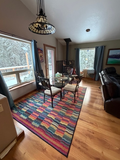 A dining area featuring a glass-topped table and chairs positioned over a vibrant, multi-colored striped rug on hardwood floors. Large windows with dark blue curtains provide a view of a snowy outdoor scene, and a wood-burning stove stands in the corner of the room.