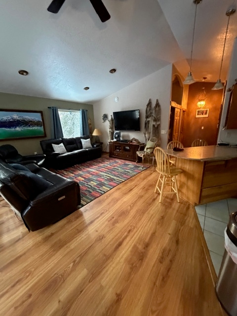 A spacious living area with hardwood floors featuring black leather sofas arranged on a colorful, multi-colored striped area rug. A flat-screen television is mounted on the far wall, and a wooden breakfast bar with chairs is visible to the right.