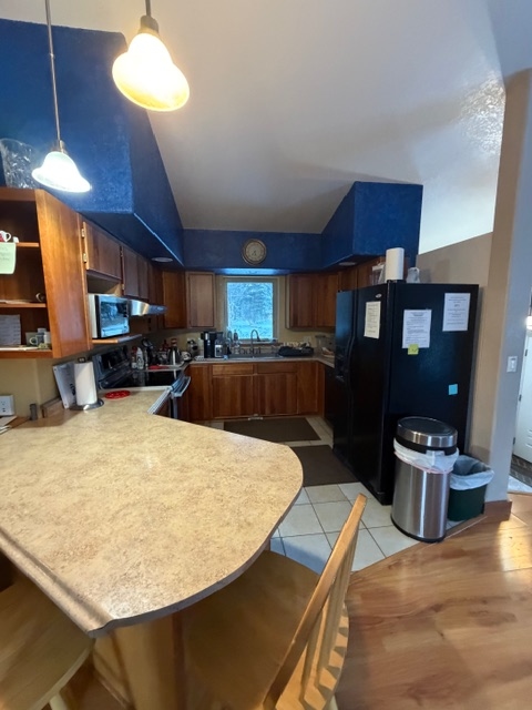 A modern kitchen featuring light-colored countertops, wooden cabinetry, and a black refrigerator. The space has a vaulted ceiling with dark blue accents above the cabinets, pendant lighting, and a view of the adjacent hardwood flooring and breakfast bar area.