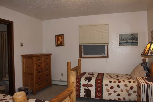 An interior view of a bedroom featuring two twin-sized log beds with nature-themed bedding. One bed is covered in a cream quilt with brown bear and red geometric patterns, while a wooden nightstand beside it holds a lamp with a forest-silhouette lampshade. The room has white walls, a large window looking out onto a dark, wintry landscape with evergreen trees, and a wooden dresser in the background.