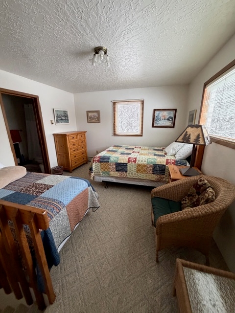 An interior view of a bedroom featuring two beds. The bed in the background is covered in a colorful block-patterned patchwork quilt, while the bed in the foreground has a blue and brown patterned quilt. The room includes a wicker armchair, a wooden dresser, and a bedside lamp with a forest-themed shade. A window offers a view of a snowy outdoor scene, and several framed pictures are mounted on the white walls.