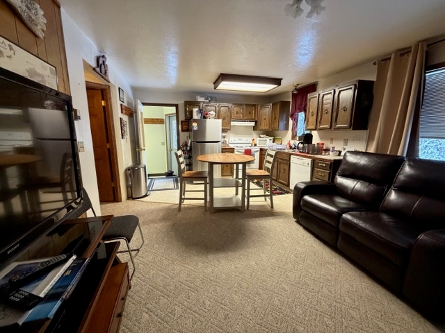 An interior view of a multi-purpose living space featuring a dark leather sofa in the foreground and a small wooden dining table with two chairs in the center. In the background, there is a compact kitchen with dark wood cabinets, a white stove, and a stainless steel refrigerator, alongside a doorway leading to another room.