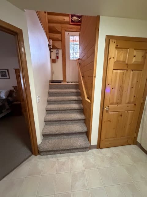 A carpeted staircase with a wooden handrail leads up toward a doorway and a light-filled upper level in a rustic interior. The space features light-colored floor tiles in the foreground and natural wood doors and wall paneling.