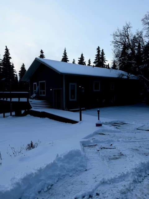 An exterior view of a long, dark-colored building with a snow-covered gabled roof in a winter landscape. A wooden ramp leads to an entrance, and the structure is surrounded by tall evergreen trees under a pale sky.