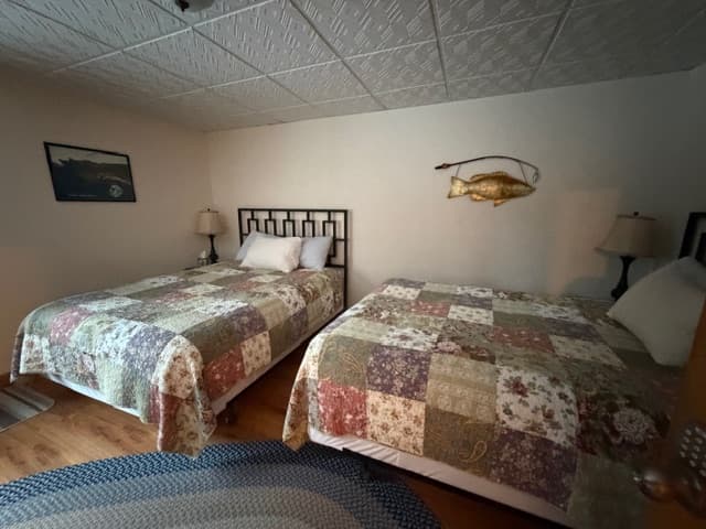 A bedroom featuring two beds with coordinated floral patchwork quilts in neutral tones of cream, tan, and brown. A large decorative golden fish mount hangs on the wall between the beds, and the room includes a white grid-patterned ceiling, bedside lamps, and a blue oval rug on the floor.