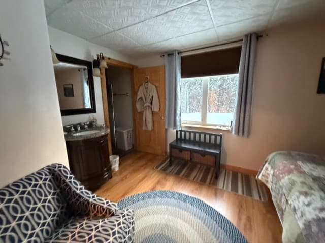 An interior view of a bedroom featuring hardwood floors and a white grid-patterned ceiling. The room includes a bed with a floral patchwork quilt on the right, a patterned armchair in the foreground, and a circular blue and white rug. A wooden bench sits under a window overlooking a snowy scene, and a dark wooden vanity with a mirror is visible on the left side of the room.