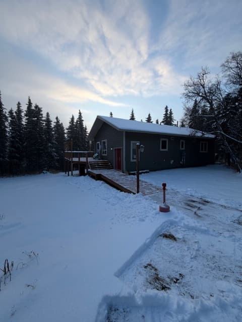 An exterior view of a one-story, dark-colored house with a snowy roof, situated in a winter landscape surrounded by tall evergreen trees under a cloudy sky. A long wooden ramp leads up to the front entrance, and the ground is covered in a thick layer of snow.