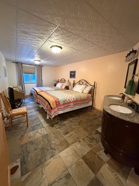 A bedroom featuring two beds with coordinated patchwork quilts in shades of peach, yellow, and blue. The room has large stone-patterned floor tiles, a white grid-patterned ceiling, and a dark wood double vanity with white sinks on the right. In the background, a window with neutral curtains looks out onto a snowy outdoor scene.