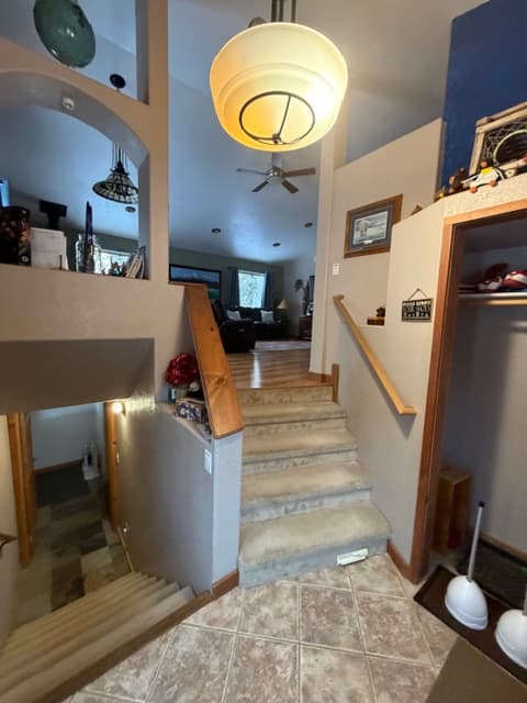 An interior entryway featuring carpeted stairs leading up to a main living area and tiled stairs leading down to a lower level. The space is illuminated by a large, round pendant light and includes a wooden handrail, neutral-toned walls, and arched cutouts overlooking the stairs.