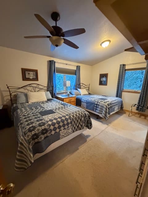 A spacious bedroom with vaulted ceilings and a ceiling fan, featuring two beds with blue and white patterned quilts. The room has neutral carpeting, blue curtains on two windows, and several framed pictures on the walls.