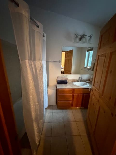 A simple bathroom featuring a light-colored tiled floor, a wooden vanity with a white sink and mirror, and a white shower curtain partially visible on the left.
