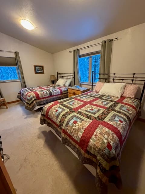 A bedroom featuring two beds with coordinated lodge-style patchwork quilts in shades of red, green, and cream. The room has neutral carpeting, vaulted ceilings, and windows framed by simple gray curtains that look out onto a snowy wooded landscape.
