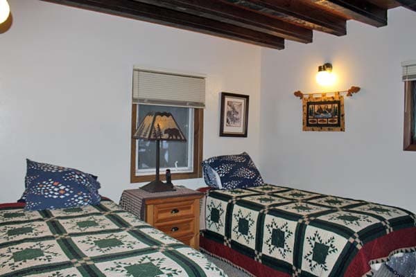 An interior view of a bedroom featuring two twin beds with matching green and cream star-patterned quilts. Between the beds is a wooden nightstand topped with a decorative lamp featuring a bear silhouette on the shade. The room has dark wood ceiling beams, white walls, and a small window overlooking a snowy scene.