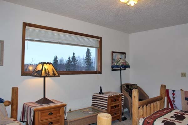 An interior view of a bedroom featuring two twin beds covered in green, cream, and red star-patterned quilts. Between the beds is a wooden nightstand with a lamp featuring a silhouette of a bear on its shade, and above one bed hangs a decorative rustic sign with a lantern. The room has white walls, dark exposed ceiling beams, and a small window overlooking a wintry outdoor scene.