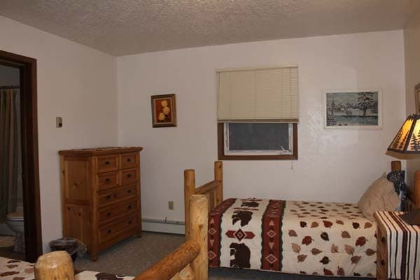 An interior view of a bedroom featuring two twin-sized log beds with nature-themed bedding. One bed is covered in a cream quilt with brown bear and red geometric patterns, while a wooden nightstand beside it holds a lamp with a forest-silhouette lampshade. The room has white walls, a large window looking out onto a dark, wintry landscape with evergreen trees, and a wooden dresser in the background.