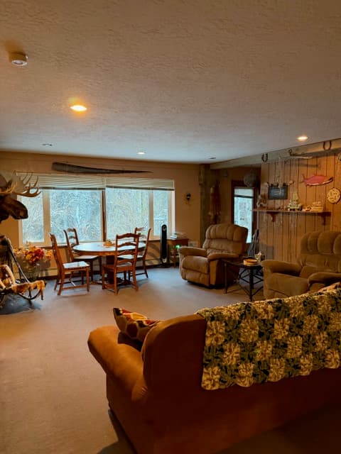 A spacious living area featuring a brown sofa draped with a green and white patterned blanket in the foreground, leading to a wooden dining set near a large window and comfortable recliners against a wood-paneled wall.