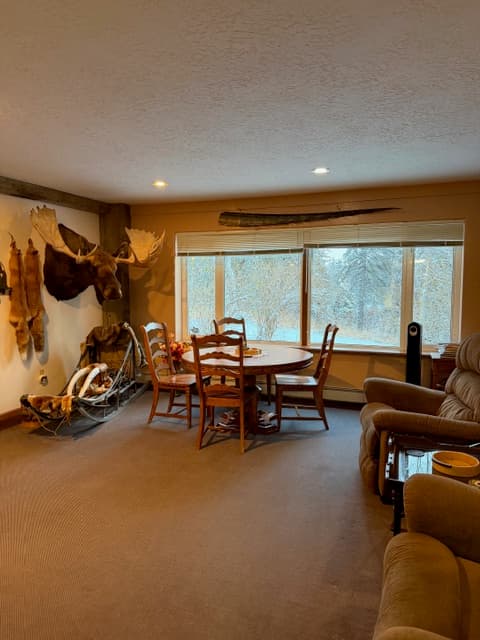 A brightly lit dining area featuring a round wooden table with four chairs set in front of a large window. To the left, a decorative wooden dog sled sits beneath a large moose antler mount on a white wall. The room has neutral-colored carpeting and recessed ceiling lights.