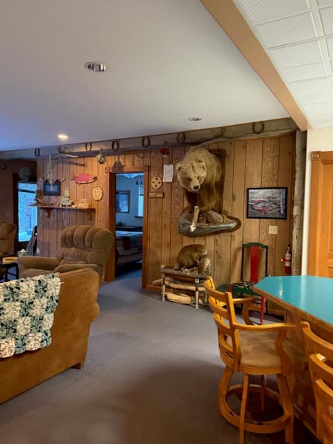 A spacious living area featuring wood-paneled walls decorated with taxidermy, including a large bear mount and a smaller animal mount below it. In the foreground, a teal-colored countertop with wooden barstools is visible, while the background shows a brown sofa and a doorway leading to another room.