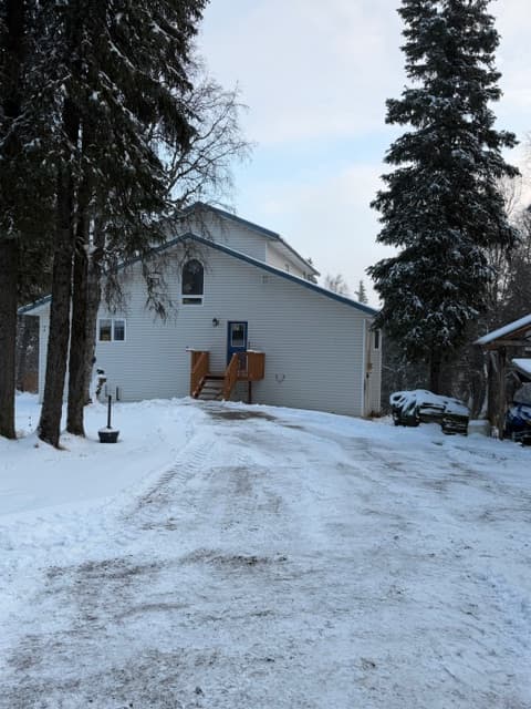 An exterior view of a white, two-story building in a snowy landscape, surrounded by tall, frost-covered evergreen trees. A small wooden deck with stairs leads to an entry door on the side of the house.