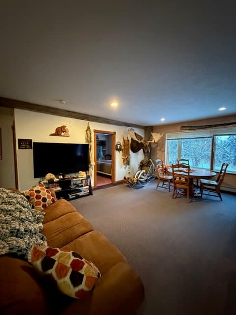 A spacious living area featuring a large flat-screen television on the left and a round wooden dining table by a bright window on the right. The walls are decorated with various items, including a moose antler mount and a decorative dog sled, with a doorway in the center leading to another room.