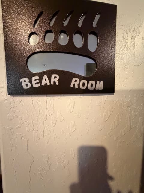 A close-up of a metal sign shaped like a bear's paw print with the words "BEAR ROOM" cut out of the bottom. The sign is mounted on a textured white wall.