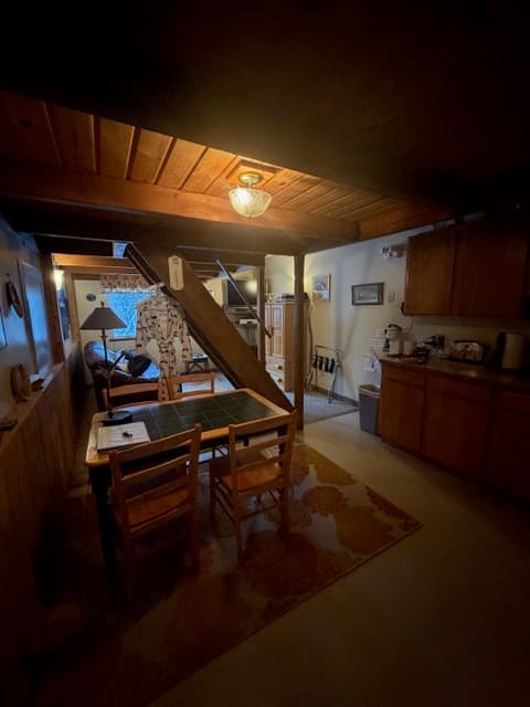 A dimly lit interior space with wood-beamed ceilings and wood-paneled walls. In the foreground, a small wooden table with two chairs sits on a patterned rug. To the right, a kitchen area with dark wooden cabinetry is visible. In the background, a staircase leads upward, and a peek into a living area reveals a lamp and a window.