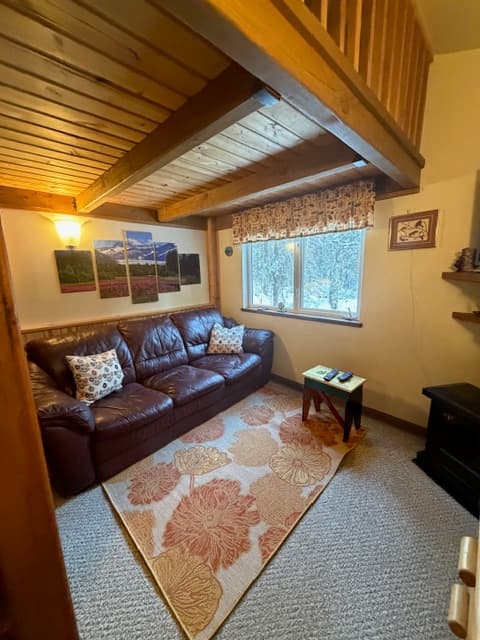 A cozy living area featuring a brown leather sofa with two patterned pillows, situated beneath a wooden loft ceiling with exposed beams. The room includes a large window with a floral valance, a floral-patterned area rug on gray carpeting, and a multi-panel mountain landscape artwork on the wall above the sofa.