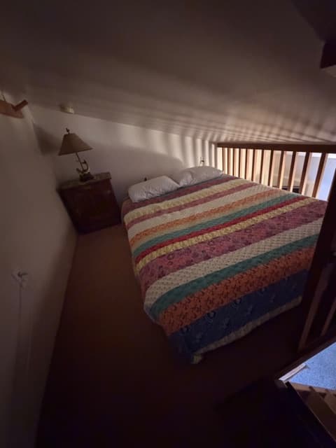 A loft bedroom with a sloped white ceiling featuring a bed with a colorful striped quilt. A wooden nightstand with a lamp sits in the corner, and a wooden railing overlooks the floor below.