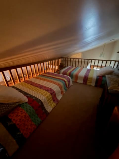 A loft bedroom with a sloped white ceiling featuring two twin beds, each covered with a colorful striped quilt. A wooden railing runs along the edge of the loft, overlooking the room below.