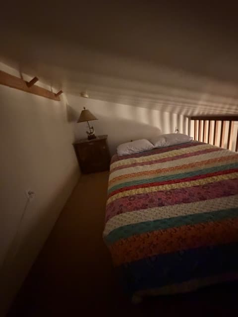 A loft bedroom with a low, sloped white ceiling features a bed covered in a colorful striped quilt. To the left of the bed stands a dark wooden nightstand topped with a lamp, and a wooden railing runs along the right side of the space.