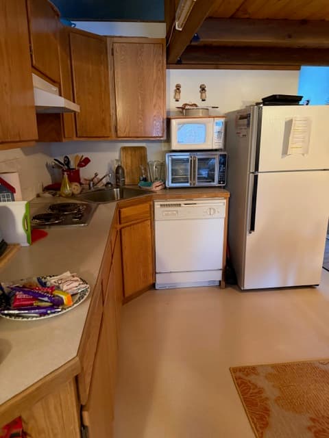 A compact kitchen area featuring wooden cabinetry, a white dishwasher, and a white refrigerator. A small white microwave and a toaster oven sit on the counter next to the refrigerator, while a plate of snacks is visible on the foreground counter. The space is partially situated beneath a wooden beamed ceiling.