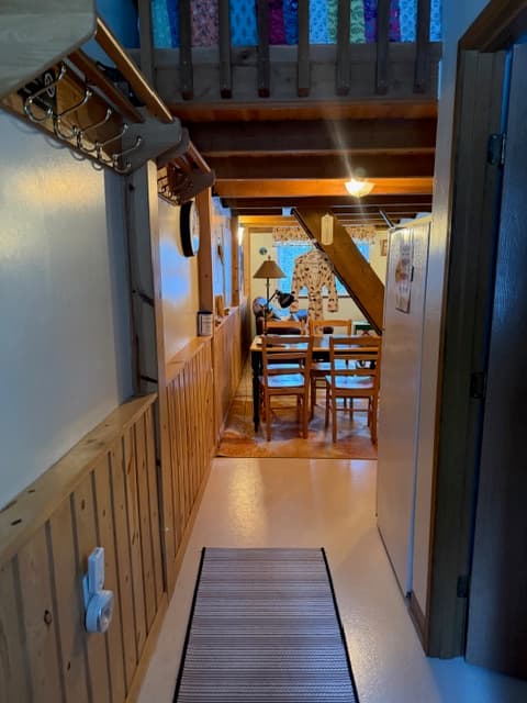 A narrow hallway with a light-colored floor runner leads into a dining area with a wooden table and chairs. The space features wood-paneled walls on the left and a white refrigerator on the right, all situated beneath a rustic wooden loft and beamed ceiling.