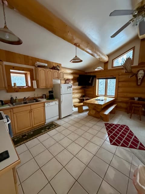 A spacious log-cabin style kitchen and dining area featuring light-tiled flooring and exposed wooden log beams. The kitchen on the left has wooden cabinetry and white appliances, while the dining area on the right includes a large wooden picnic-style table with benches and a wall-mounted moose head decoration.