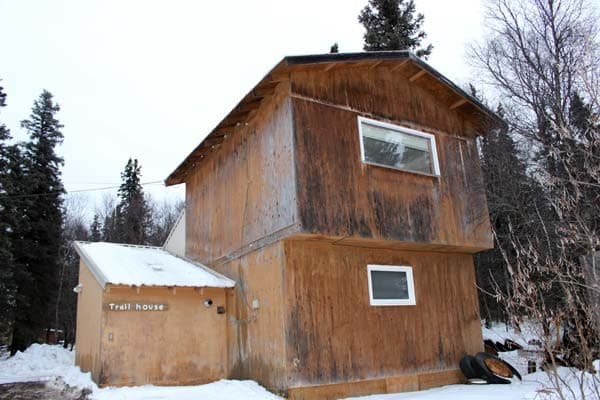 An exterior view of a two-story wooden building with a snow-covered roof, set against a backdrop of tall evergreen trees. The building features weathered wood siding and small rectangular windows, with a smaller attached structure on the left side.