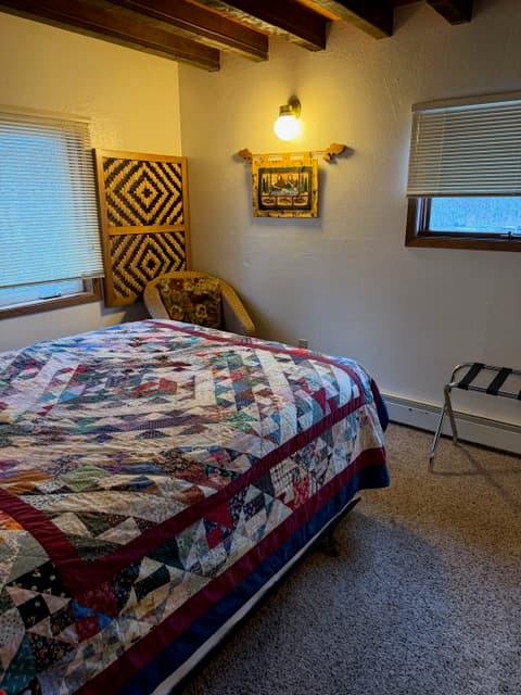 An interior view of a bedroom featuring a bed with a vibrant star-patterned patchwork quilt in shades of blue, red, and pink. The room includes a dark metal headboard, dark exposed wooden ceiling beams, and a large mirrored closet that reflects the bed and a portion of the room's white walls and tan carpeting.