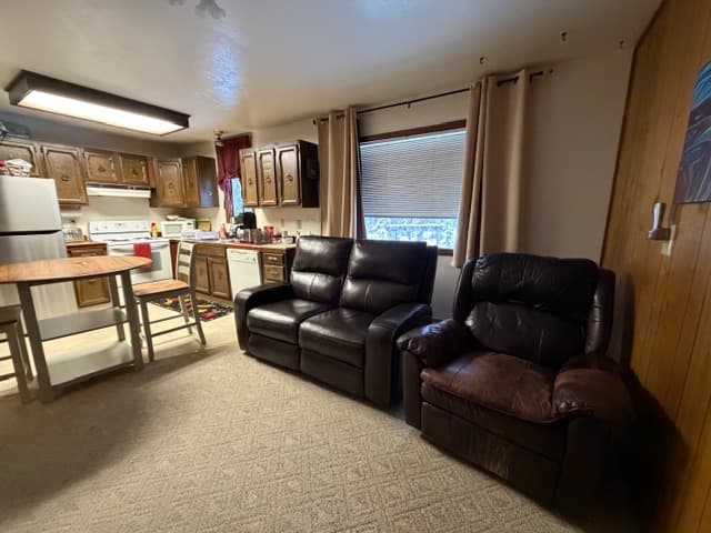 An interior view of a bedroom featuring a single bed covered in a vibrant star-patterned patchwork quilt in shades of red, blue, pink, and white. The room includes a dark metal headboard, a wicker armchair in the corner, and a small folding luggage rack. Dark exposed wooden beams run across the ceiling, and a single light fixture is mounted on the wall above the bed.