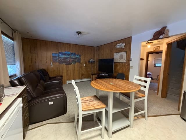 An interior view of an open-plan living and kitchen area featuring a dark leather reclining sofa and a matching armchair on a neutral-colored carpet. The space includes a small, round wooden dining table with two white chairs, wood-paneled walls decorated with rustic signs, and a television on a stand in the corner. In the background, a compact kitchen with dark wood cabinets and a white refrigerator is visible next to a doorway leading to a hallway.