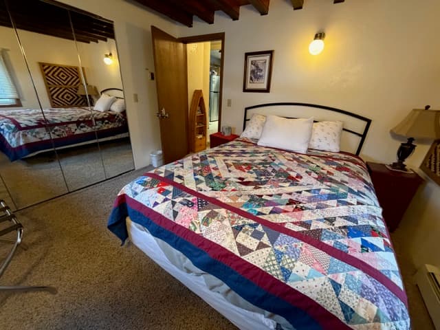 An interior view of a bedroom featuring a bed covered in a colorful star-patterned patchwork quilt with blue, red, pink, and white tones. The room includes a dark metal headboard, a large mirrored closet reflecting the bed, and dark exposed wooden ceiling beams. A doorway in the background reveals a glimpse of a hallway.